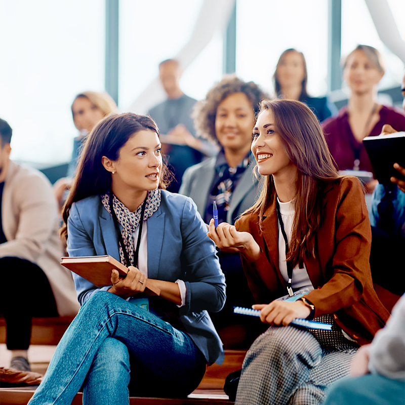Two women sit and talk while holding notebooks in a bright, modern lecture hall filled with other people. The group appears engaged and attentive, suggesting a professional or educational setting.