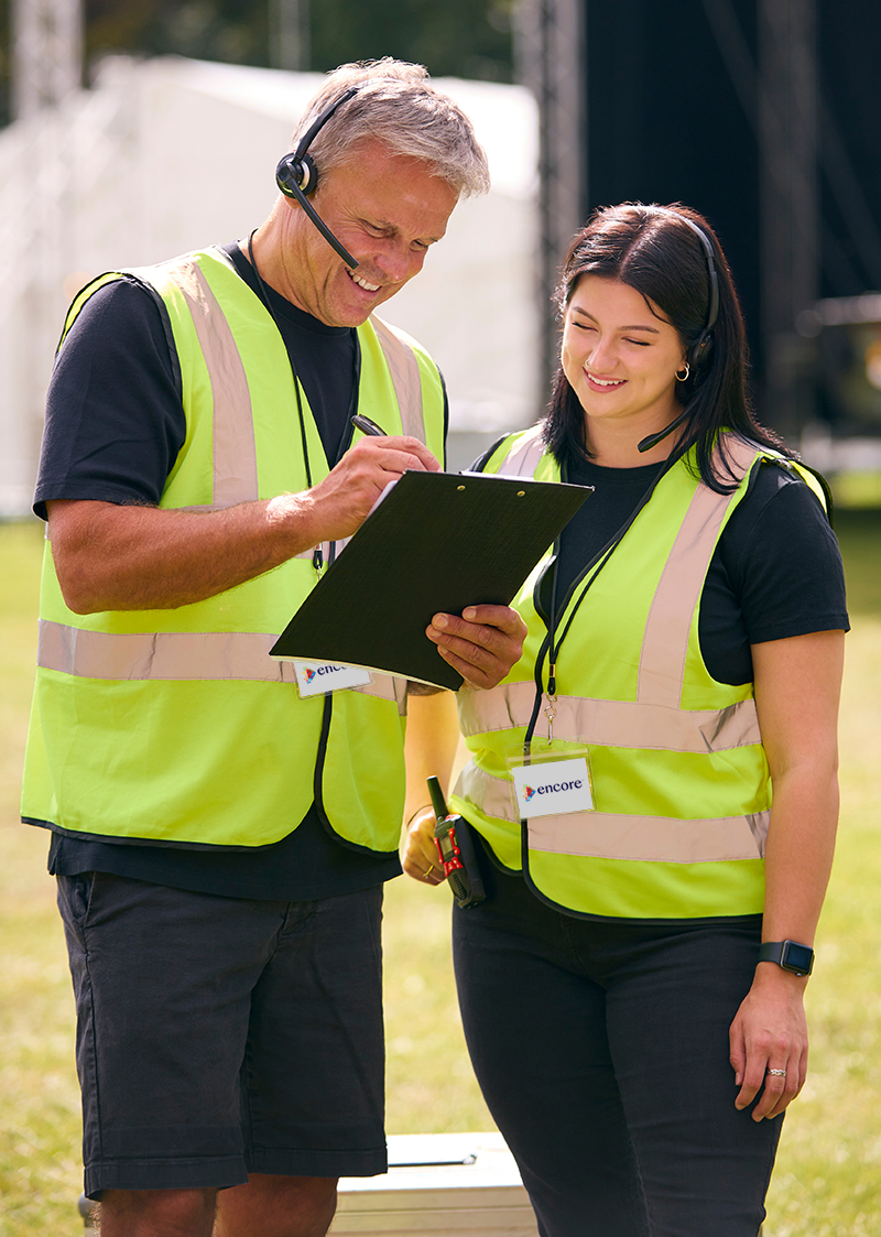 Two event staff in yellow safety vests and headsets stand outdoors, smiling as one writes on a clipboard while the other looks on. Both wear black shirts and name badges.
