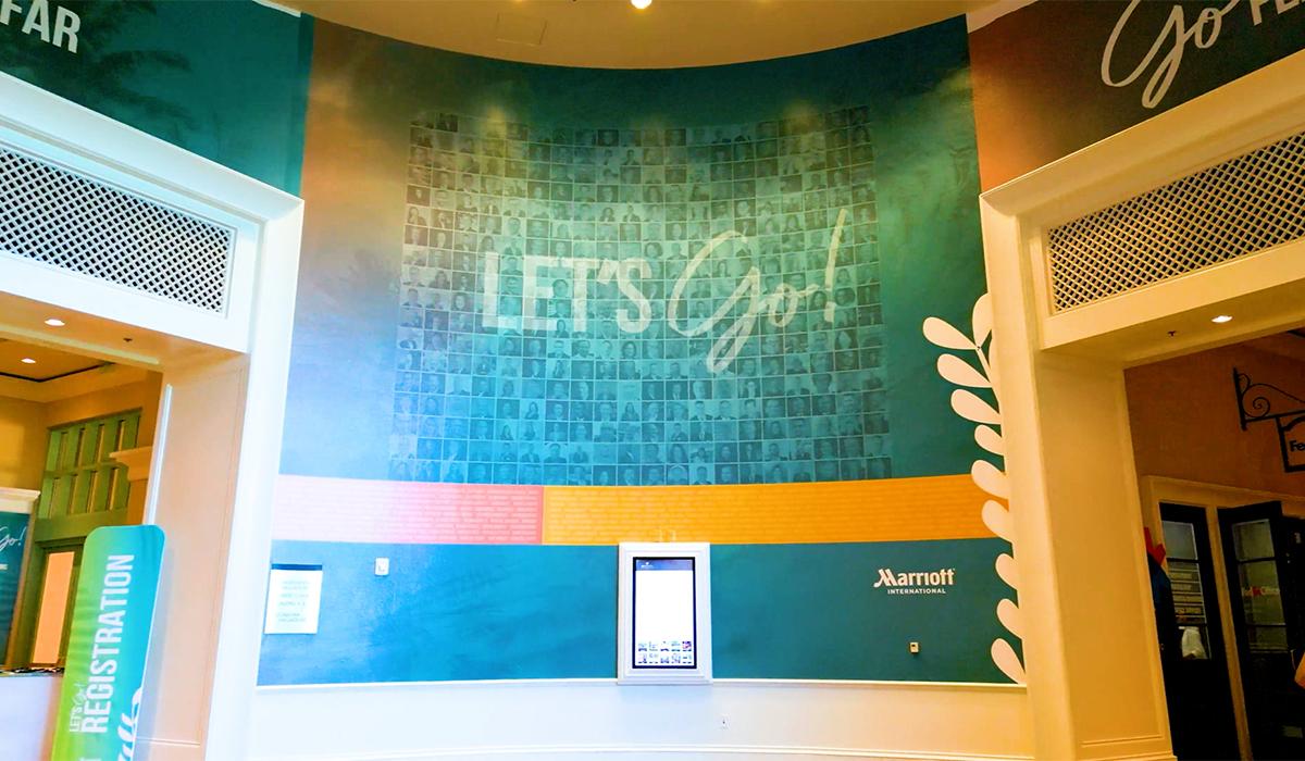 Large teal wall inside a Marriott conference venue features a mosaic of small black-and-white portraits and the words “LET’S Go!” in bold, bright letters. A digital kiosk stands below the mural, and a registration sign is visible on the left.