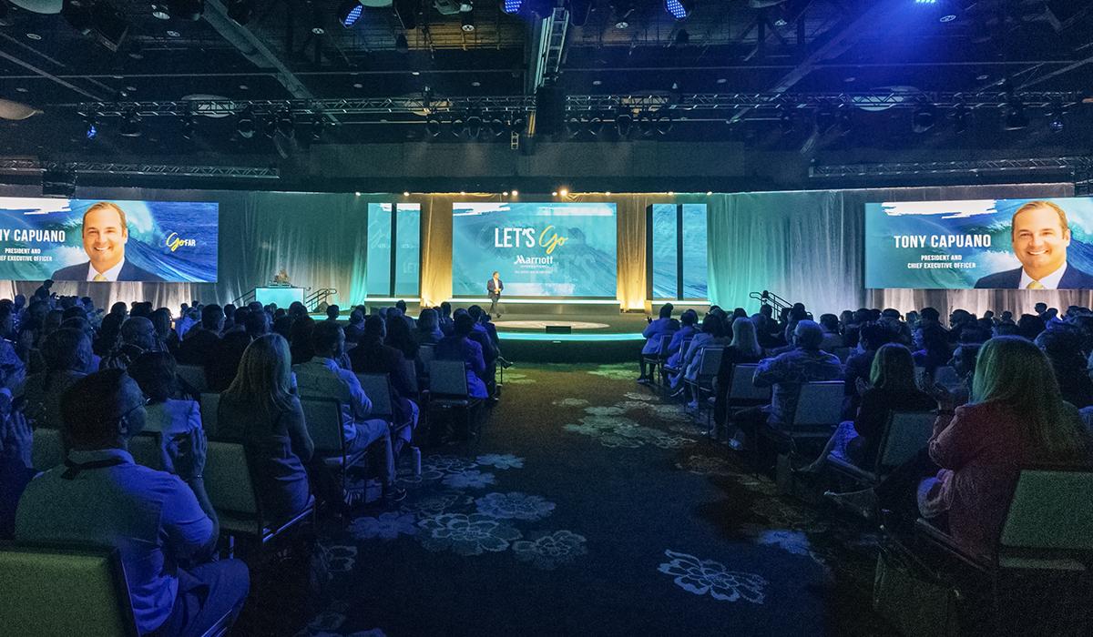 A large audience sits in a dimly lit conference hall facing a stage with a speaker. Two large screens display a mans photo and the name Tony Capuano along with the words Lets Go and Marriott.