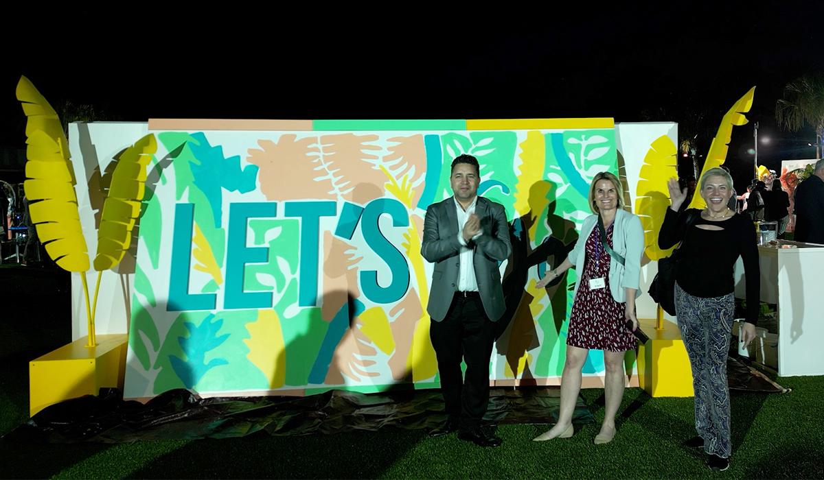 Three people stand and smile in front of a colorful, tropical-themed backdrop with large leaves and the word “LET’S” at an outdoor nighttime event. Two women and one man are posing on grass.
