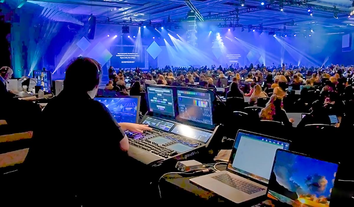 A technician operates lighting and sound equipment at the back of a large conference room filled with an audience, facing a stage illuminated by blue lights and projected graphics.