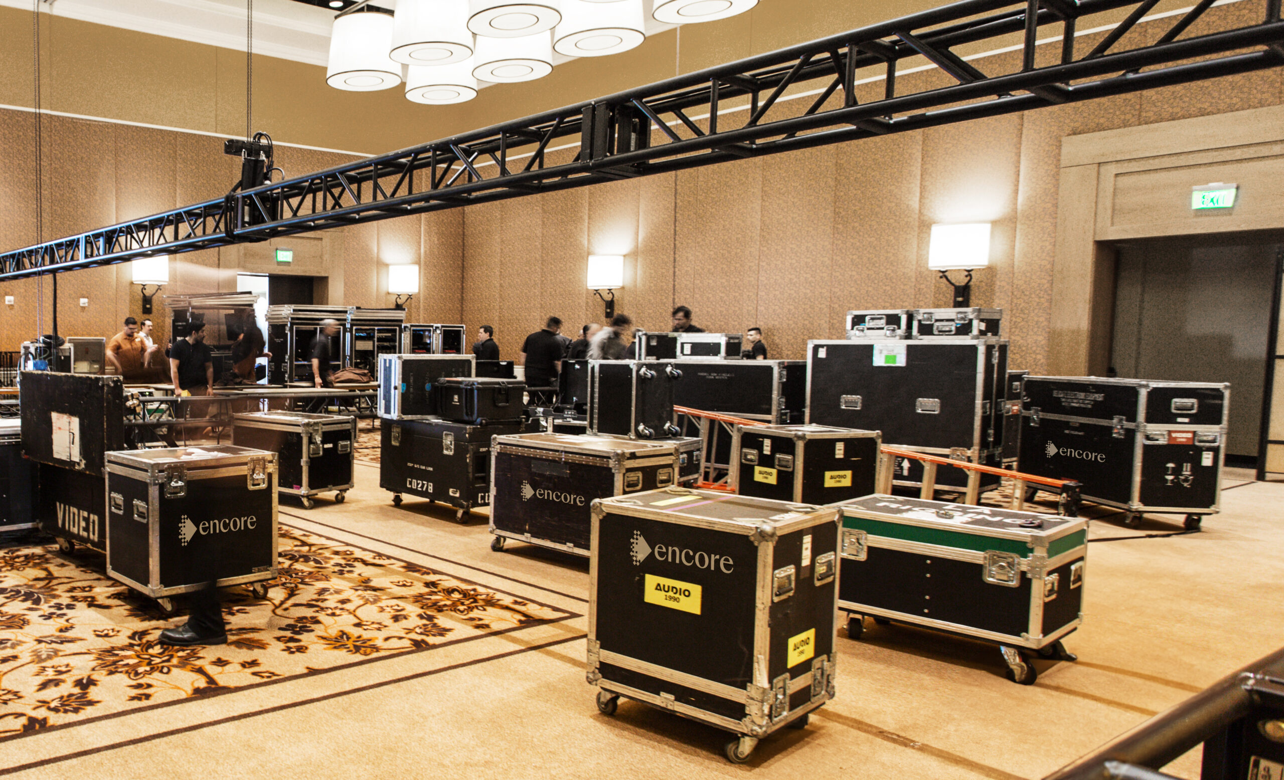Large black equipment cases are scattered around a hotel conference room. People in the background appear to be setting up audio-visual gear, with lighting trusses suspended above and patterned carpet on the floor.