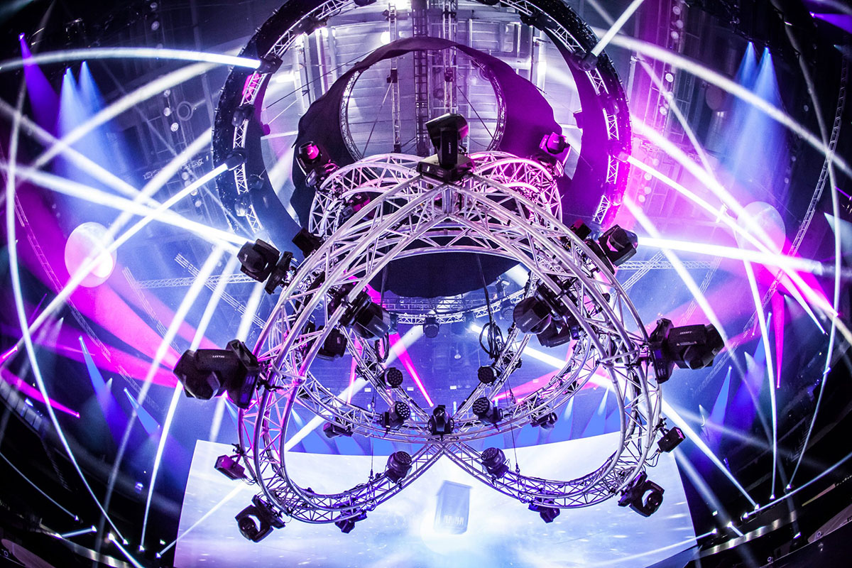 A large circular metal truss with stage lights is suspended from the ceiling, surrounded by vibrant beams of purple, white, and blue light at a concert or event venue.