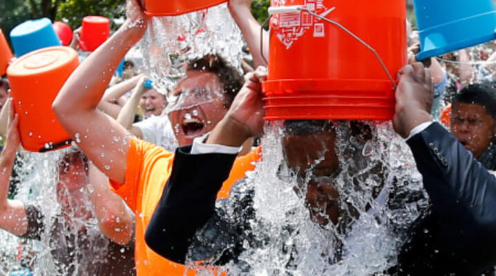 A group of people pour buckets of ice water over their heads outdoors, participating in the ALS Ice Bucket Challenge. Water splashes down as they react to the cold.