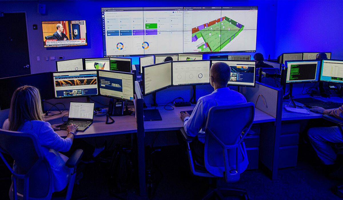Several people work at multiple computer monitors in a dimly lit control room, with large screens on the wall displaying data dashboards, maps, and news broadcasts. The room is illuminated by blue lighting.