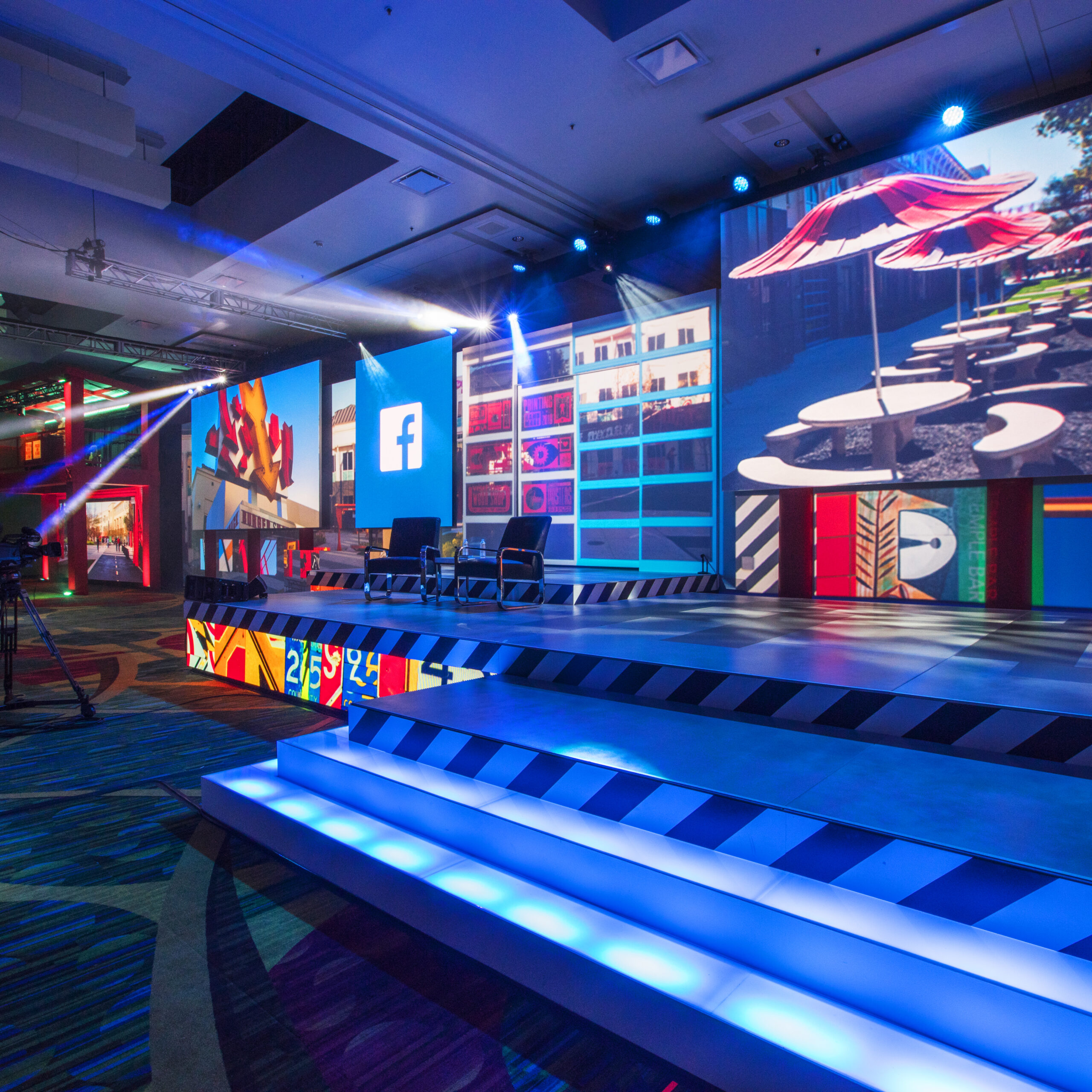 A brightly lit stage with large digital screens displaying colorful graphics and a Facebook logo, empty chairs on the stage, and glowing blue steps in a modern event venue.