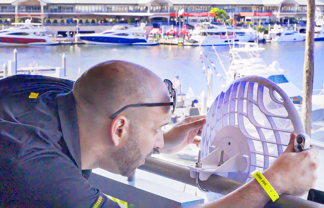 A man adjusts a wireless satellite dish on a balcony overlooking a marina with several docked yachts and boats in the background.