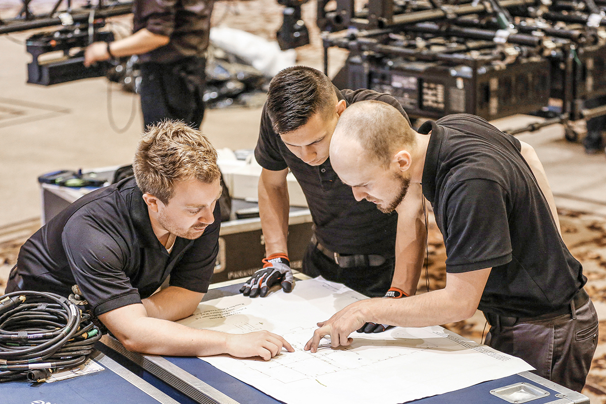 Three men in black shirts intently examine and discuss a technical blueprint or floor plan on a table, surrounded by coiled cables and equipment in an indoor work setting.