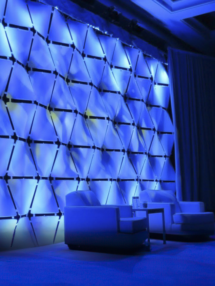 Two modern white armchairs sit beside a small table with a water bottle, in front of a glowing geometric wall lit with blue and yellow lights. A curtain hangs to the right, creating a contemporary lounge atmosphere.