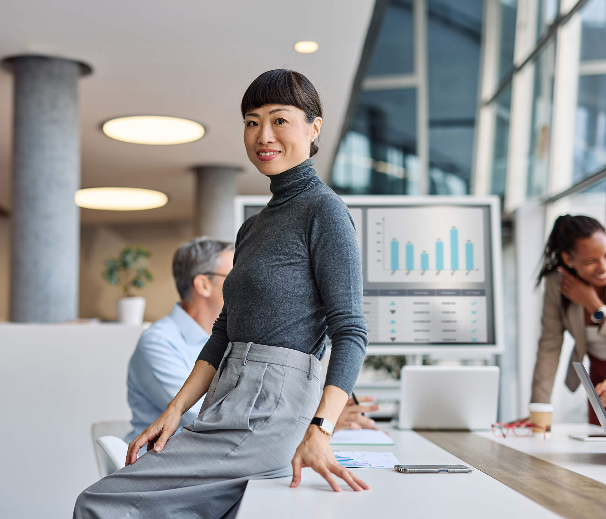 A confident woman in a gray turtleneck and trousers sits on a desk in a modern office, smiling at the camera. Two colleagues work in the background, with charts displayed on a large monitor.