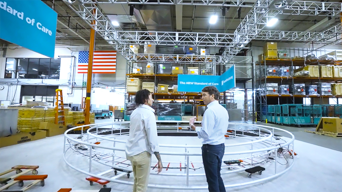 Two people stand inside a large warehouse, talking near a circular metal framework. Shelves with boxes, blue banners reading “Standard of Care,” and an American flag are visible in the background.