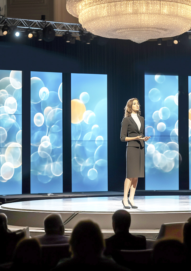 A woman in business attire stands on a round stage, presenting to an audience. Behind her, large digital screens display abstract blue and white lights. A large chandelier hangs overhead.