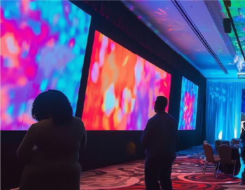 Two people stand in a dimly lit room facing large, colorful digital screens displaying abstract patterns of blue, pink, and orange; empty chairs line one side of the room.