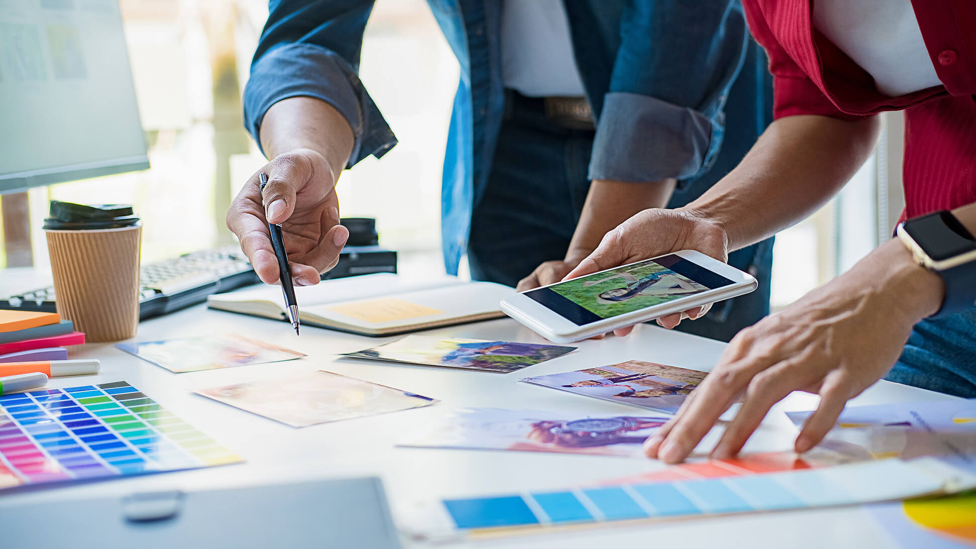 Two people review photos and color swatches on a desk, one holding a smartphone and the other pointing with a pen. Art supplies, a notebook, and a coffee cup are also visible, suggesting a creative workspace.