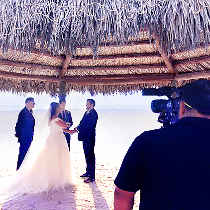 A couple stands under a thatched gazebo on a sandy beach exchanging vows, with two people beside them and a cameraman recording the ceremony.