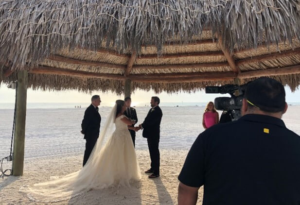 A couple stands under a thatched-roof gazebo on a sandy beach, exchanging vows during a wedding ceremony with an officiant, two guests, and a camera operator filming the scene.