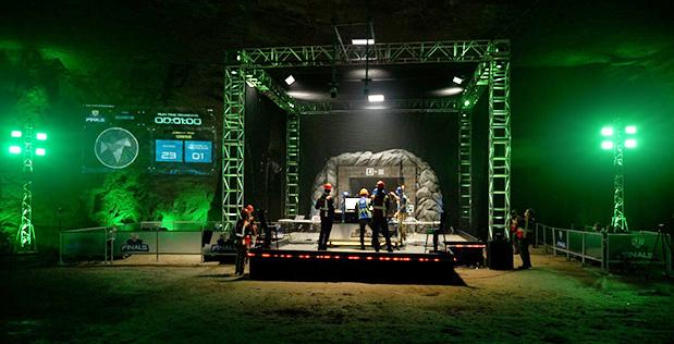 An illuminated esports stage with green lighting is set up inside a cave, featuring players and staff standing on the platform. Screens and equipment are visible, with a rock wall in the background and a scoreboard projected on the left.
