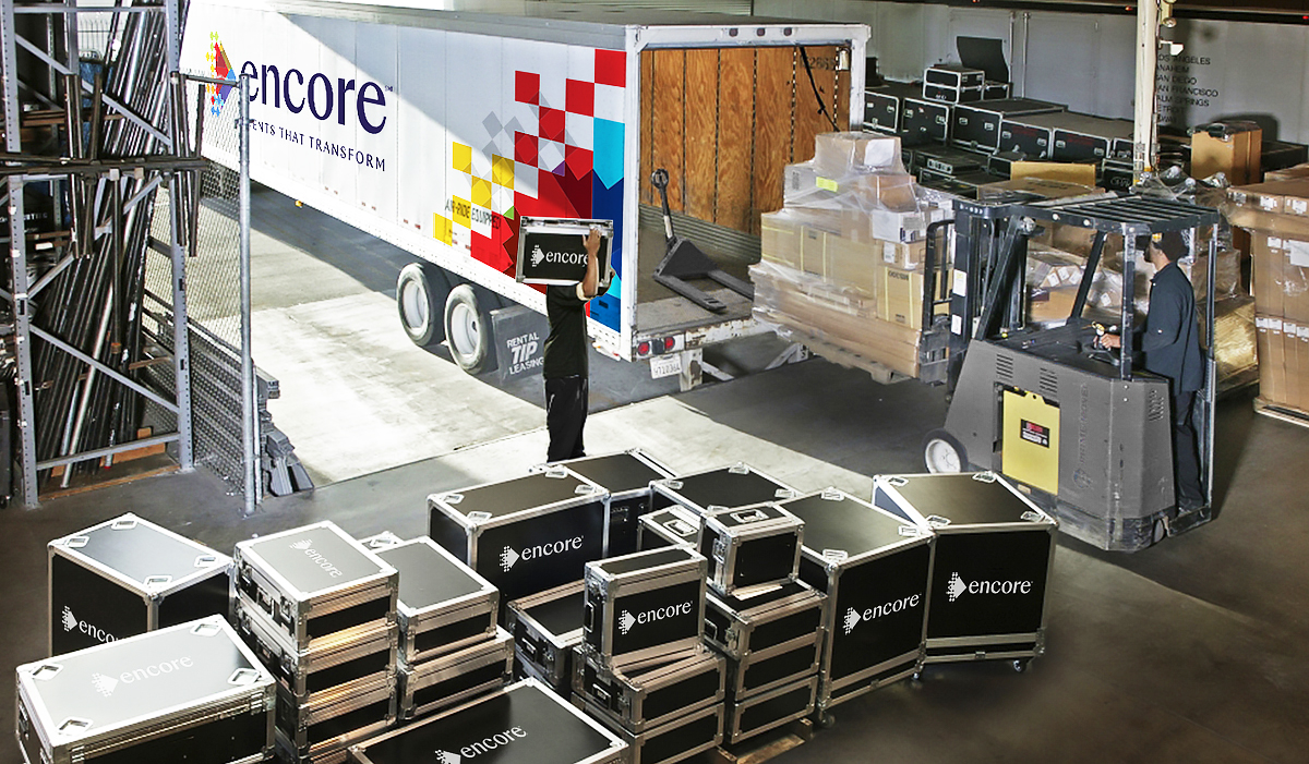 Warehouse workers load black Encore equipment cases into a truck using a forklift and by hand. The truck’s trailer is open, revealing boxes inside. More stacked cases and shelves are visible in the busy warehouse.