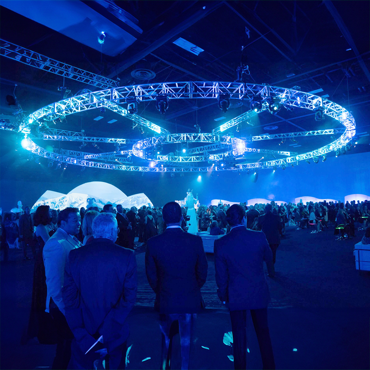 A large event space illuminated with blue lighting, featuring circular metal trusses suspended from the ceiling and a crowd of people socializing and mingling below.