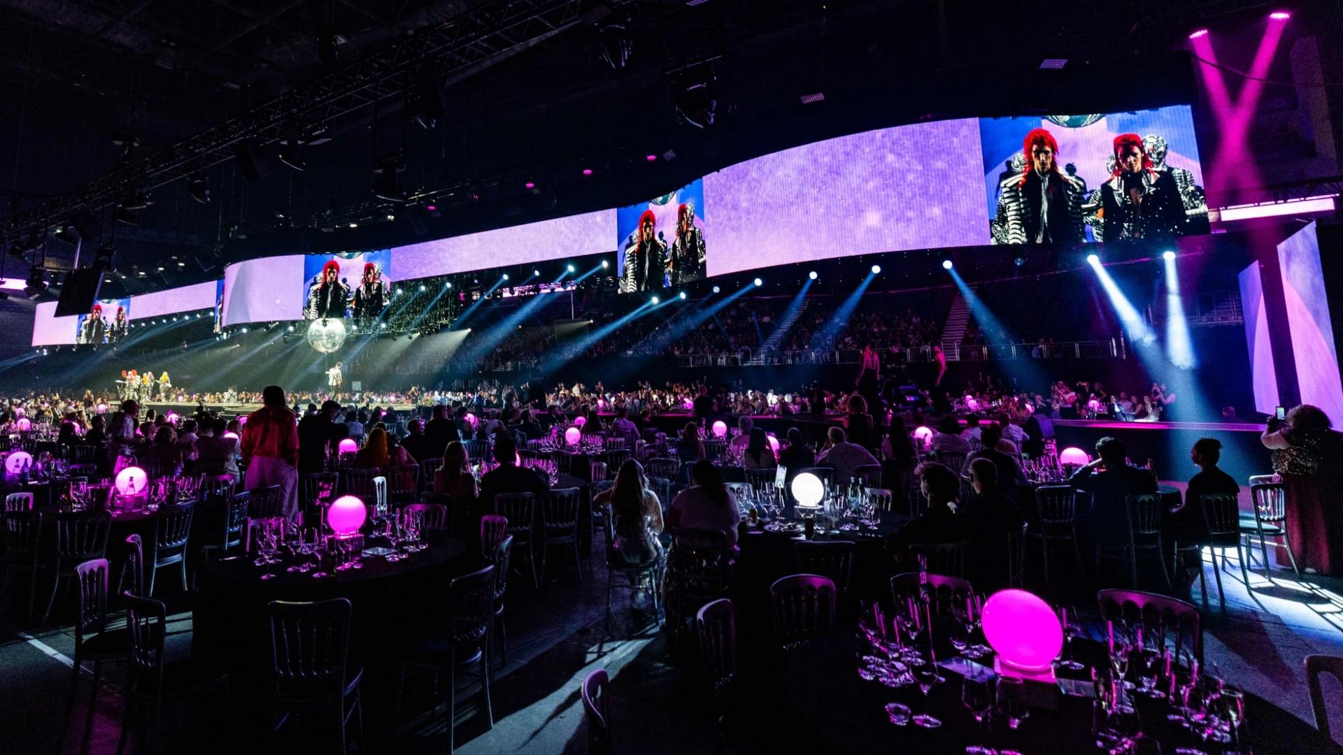 Two runway performers in sculptural costumes under dramatic arena lighting - Encore EMEA